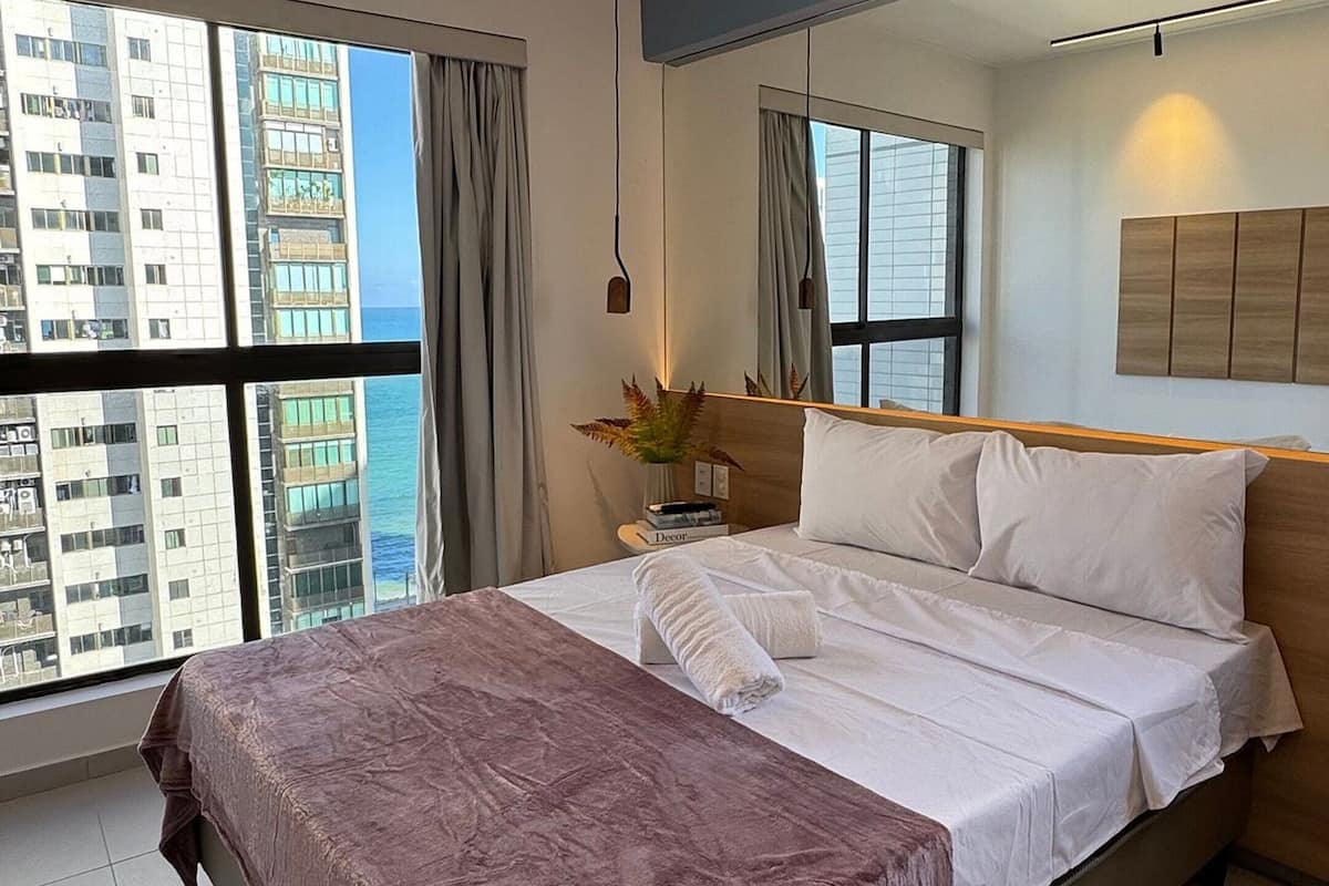 Habitación de hotel con una cama grande, una mesita de noche con un florero, vistas a la ciudad a través de ventanas amplias y un diseño de techo moderno.