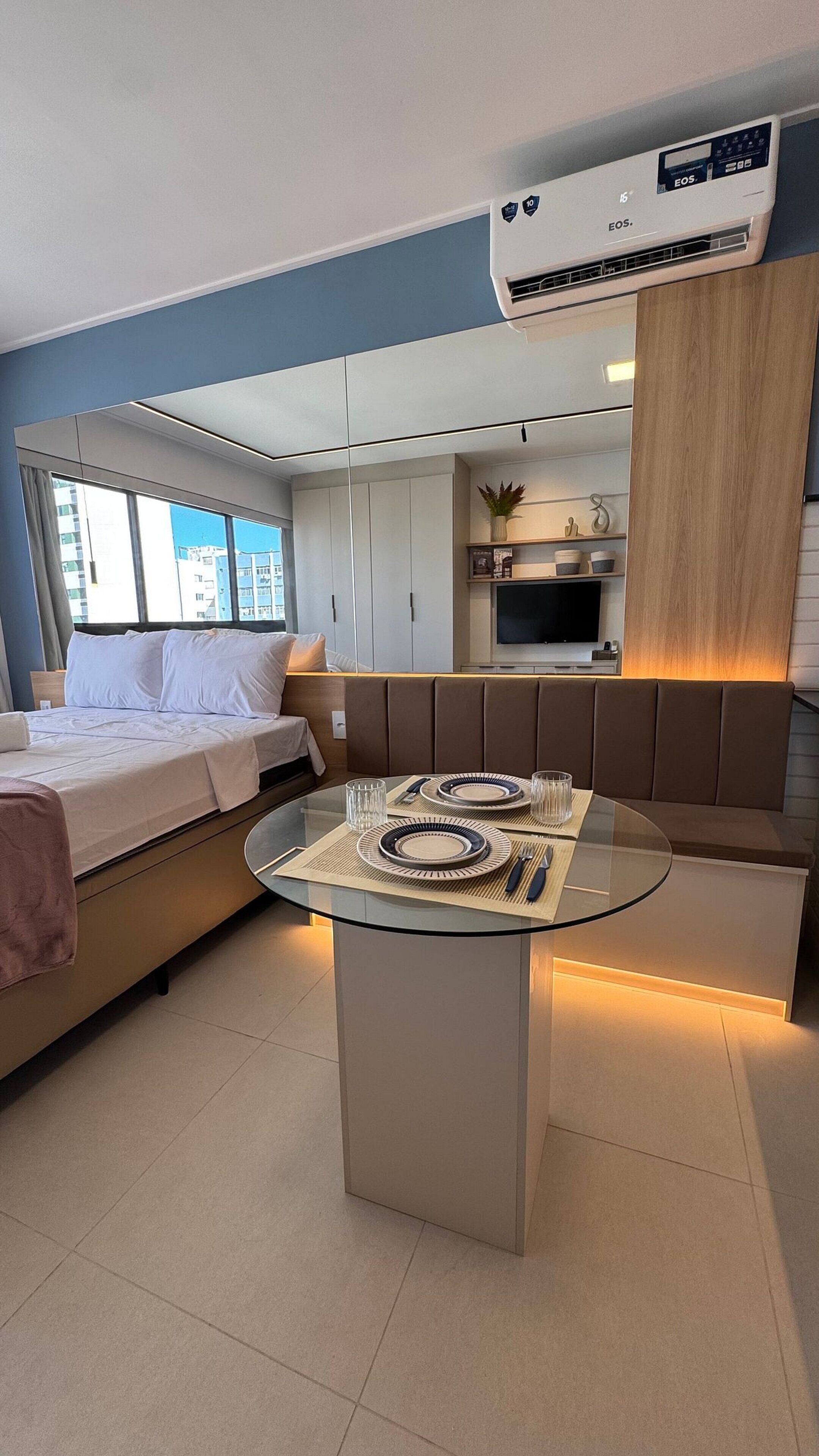 A modern hotel room with a bed, a dining table with plates and cutlery, a sofa, a TV, and an air conditioning unit.