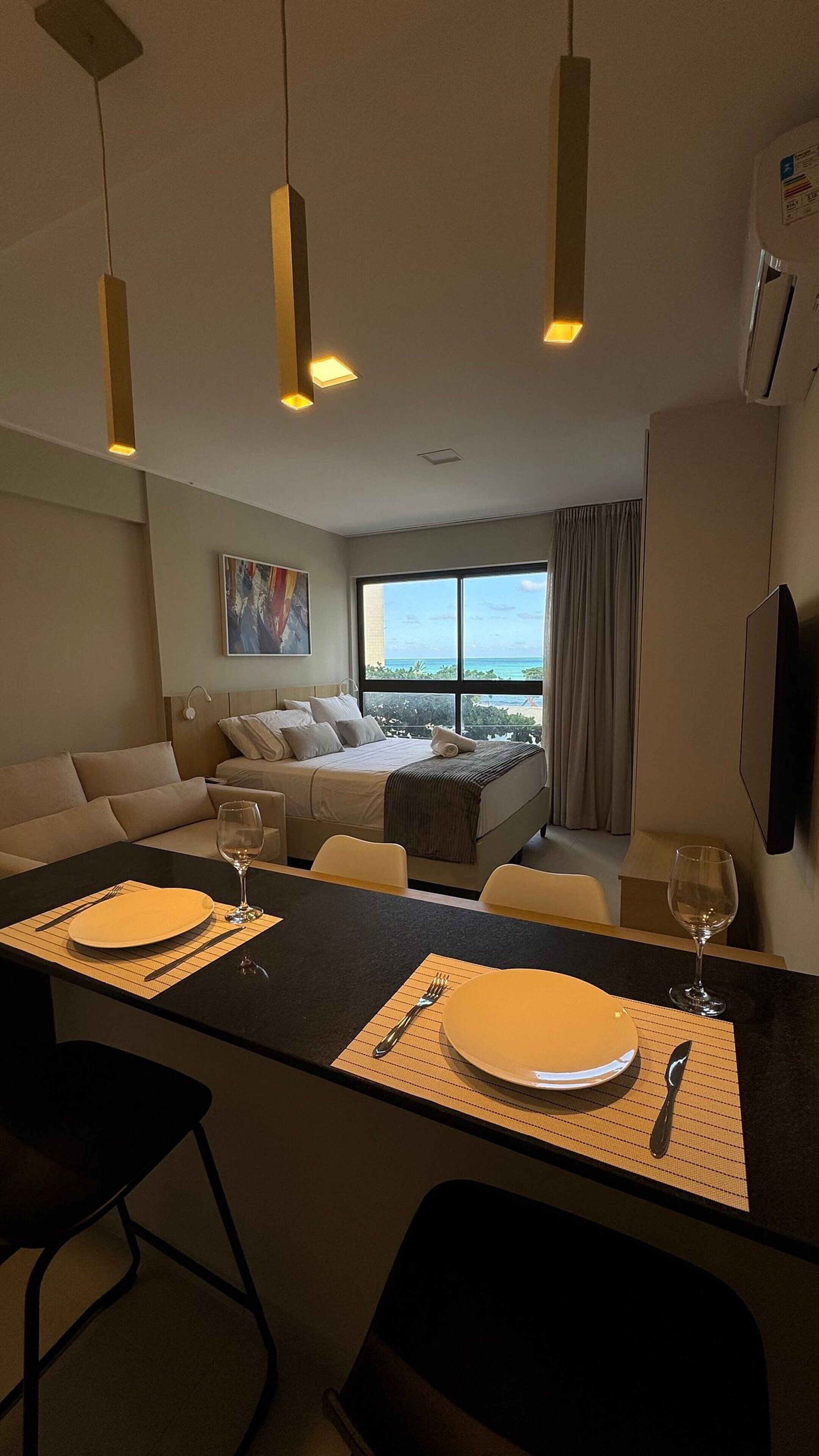 A modern hotel room with a large bed, a dining area with a table set for two, and a view of the ocean.