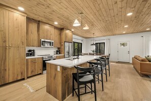 A modern kitchen with wooden cabinets, a central island, bar stools, and a dining area with a view of the outdoors. - Legend's Lodge - Yellowstone's perfect home base / AC / Wi-Fi / Fire Pit (Island Park)
