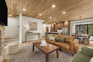 A modern living room with a wooden ceiling, a leather sofa, a coffee table, and a kitchen in the background. - Legend's Lodge - Yellowstone's perfect home base / AC / Wi-Fi / Fire Pit (Island Park)