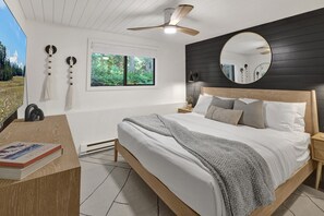 A bedroom with a bed, bedside tables, a desk, a chair, a window, a ceiling fan, and a mirror.
