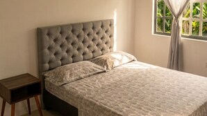 Un dormitorio con cabecera capitonada, colcha estampada, mesita de noche y ventana con cortinas.