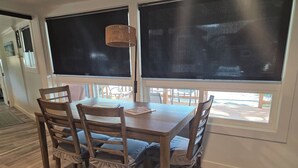 A dining area with a wooden table and chairs, large windows with blinds, and a wicker floor lamp. - Arch Cape cottage with a View (Arch Cape)