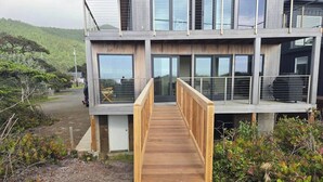 A modern two-story building with a wooden deck leading to a glass entrance. - New Modern Oceanfront W/ Panoramic Ocean Views (Rockaway Beach)