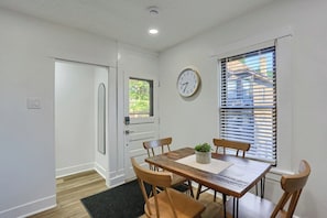 Un comedor con una mesa de madera, cuatro sillas, una planta en maceta, un reloj de pared y una ventana con persianas.