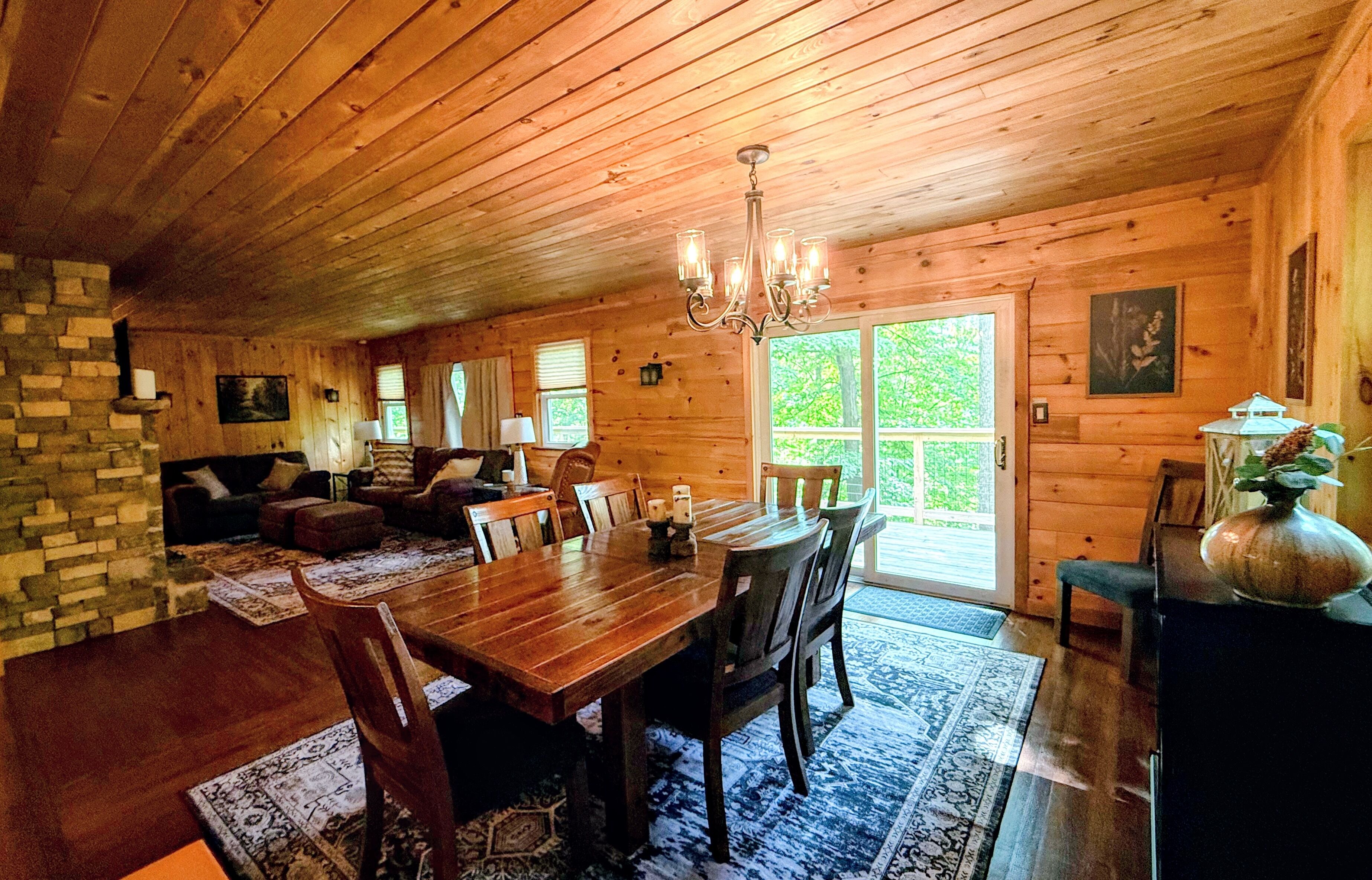 A wooden cabin interior with a dining area, living room, and a large window.
