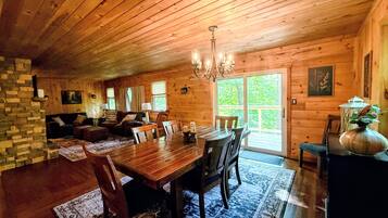A wooden cabin interior with a dining area, living room, and a large window.