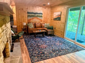 A cozy room with a stone wall, wooden paneling, a sofa, a coffee table, and a large window with a view of greenery.