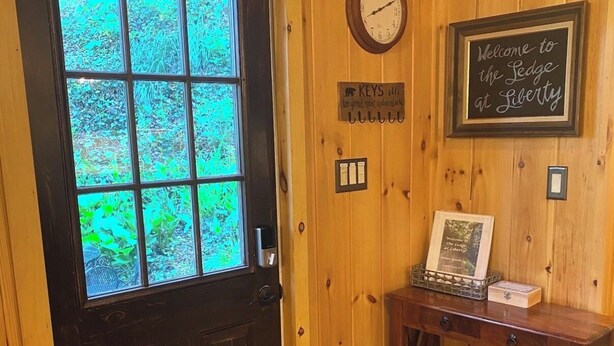 A wooden door with a window, a wall clock, a welcome sign, a small table with a framed picture, and a fire extinguisher.