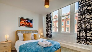 Un dormitorio con cama, mesita de noche, lámpara, cuadro enmarco, ventana con cortinas y vista a edificios.
