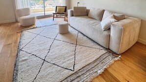 Ein modernes Wohnzimmer mit einem beige gefärbten Sofa, einem gemusterten Teppich, einem Sessel und Blick durch große Fenster.