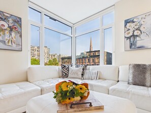 A modern living room with a white sofa, a coffee table with a floral arrangement, and large windows offering a city view.