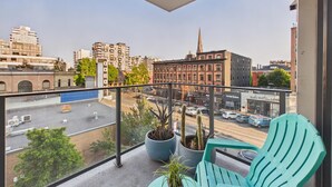 Ein Balkon mit zwei grünen Stühlen und einem kleinen Tisch, mit Blick auf eine Stadtstraße mit Gebäuden und einem Kirchturm.
