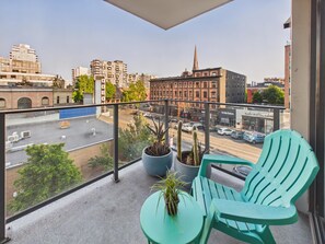 Ein Balkon mit zwei grünen Stühlen und einem kleinen Tisch, mit Blick auf eine Stadtstraße mit Gebäuden und einem Kirchturm.