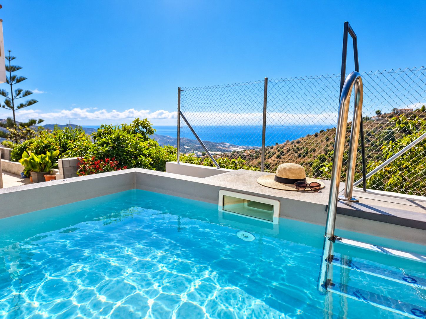 A clear blue pool with a view of the ocean, surrounded by greenery and a fence.