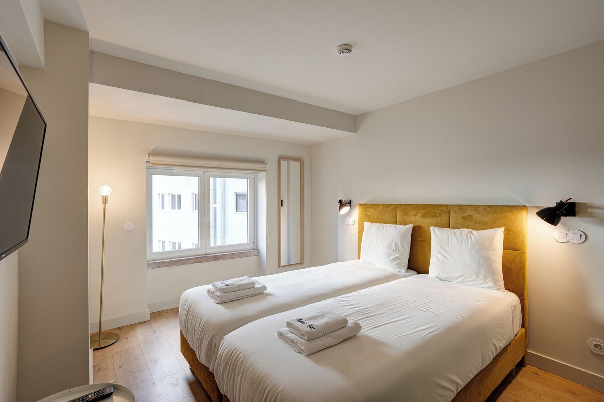 A hotel room with two beds, a TV, a floor lamp, and a window.