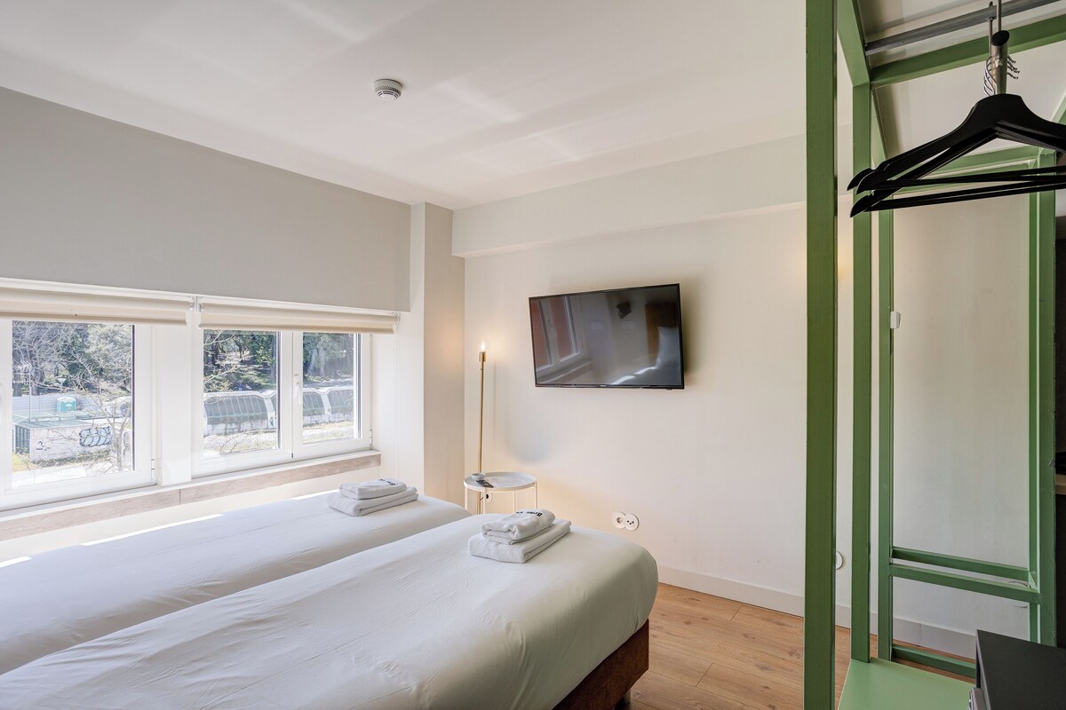 A bedroom with two beds, a TV, a hanging clothes rack, and a window with a view.