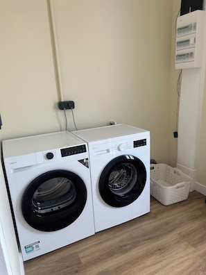 A laundry room with two white front-loading washing machines, a white laundry basket, and a wall-mounted electrical outlet.