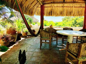 A covered outdoor seating area with a table and chairs, surrounded by potted plants and a thatched roof.