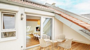 Un balcón con suelo de madera y sillas blancas, una puerta corredera de vidrio que da a un espacio interior con una mesa y sillas de comedor.