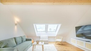 Una habitación estilo loft con techo inclinado, un sofá verde, una pequeña mesa, una silla blanca, un televisor sobre un mueble blanco y un tragaluz grande.