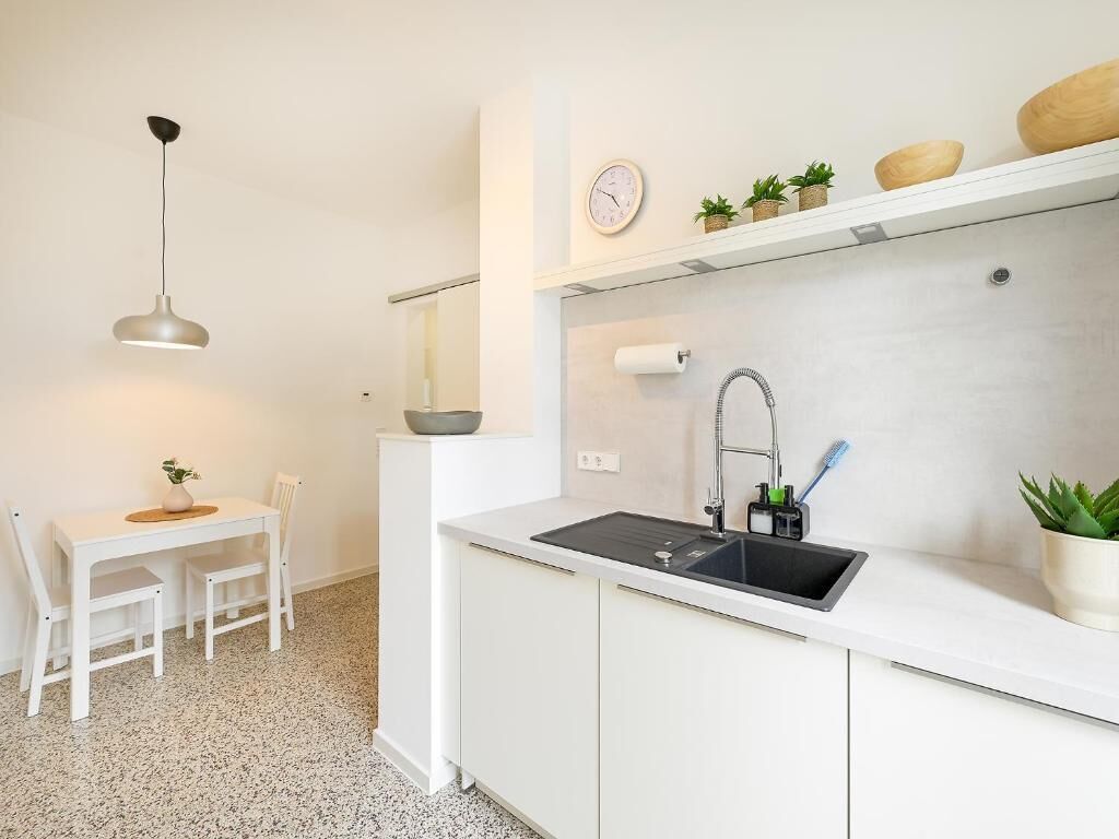 A compact kitchen with a sink, a small dining table with chairs, a pendant light, and a clock on the wall.