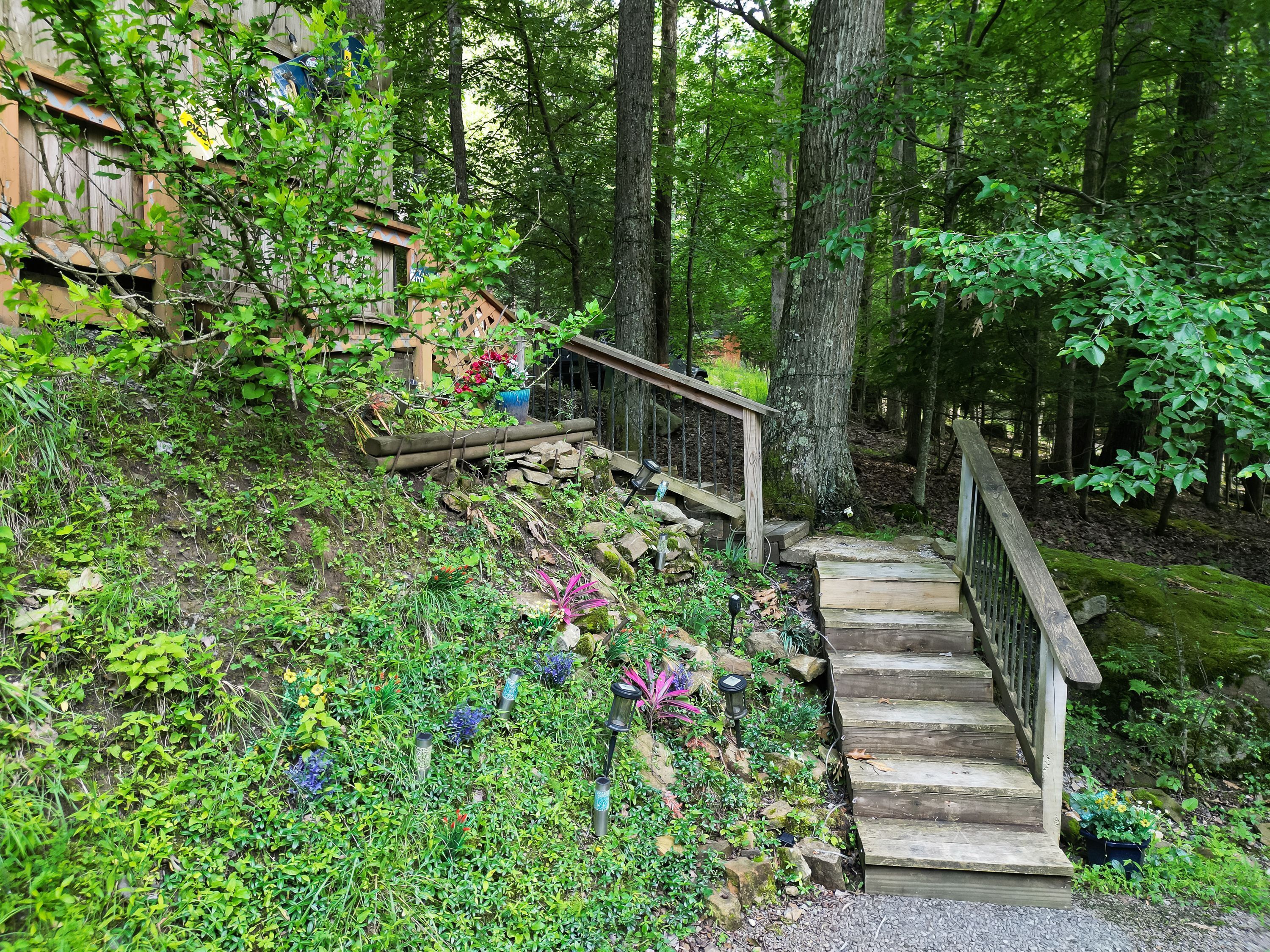 A wooden staircase leading up a grassy slope with a metal railing, surrounded by dense greenery and trees.