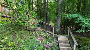 Eine hölzerne Treppe führt einen grasbewachsenen Hang hinauf, gesichert durch ein Metallgeländer, umgeben von dichtem Grün und Bäumen.