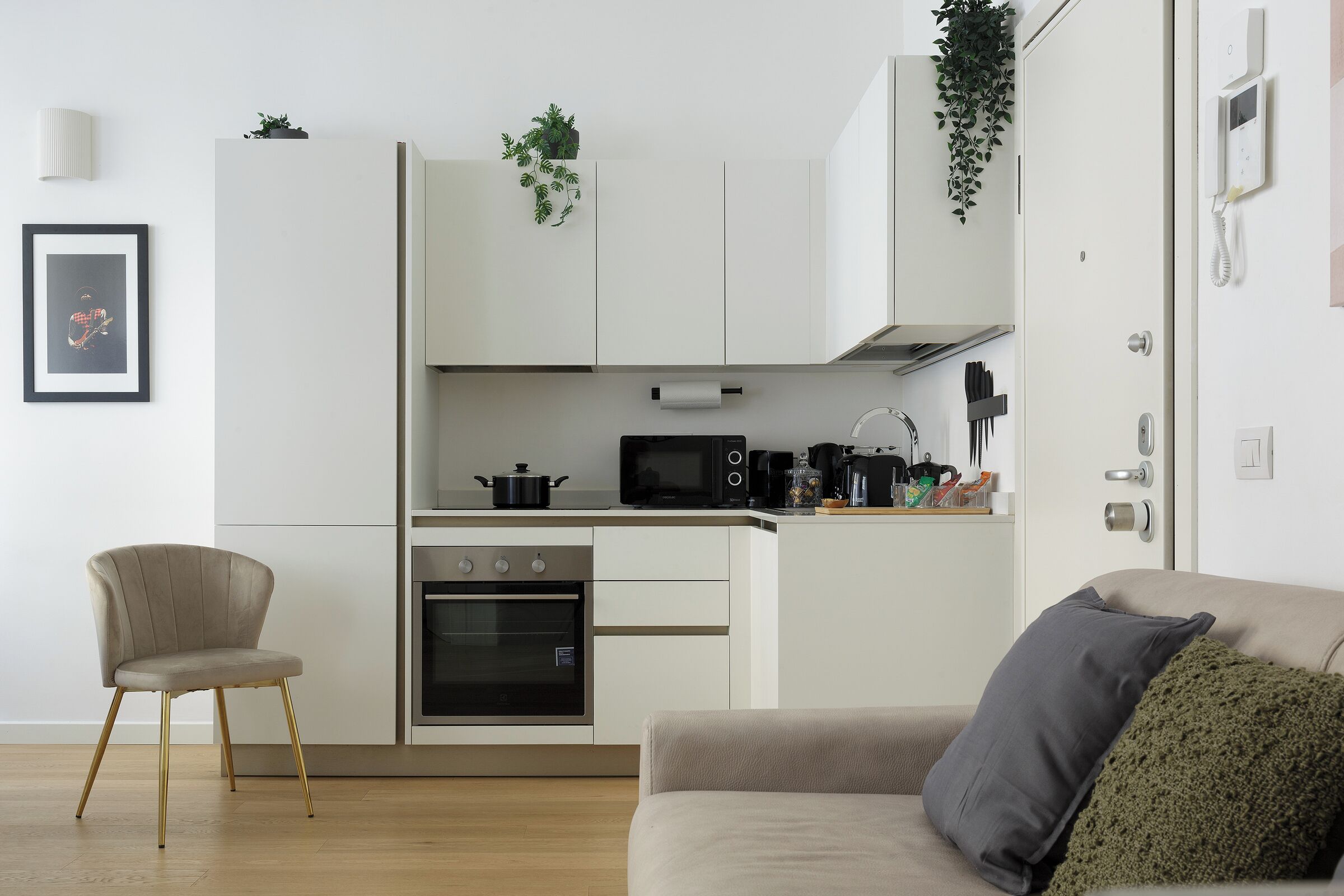 Una cucina moderna con mobili bianchi, forno integrato, microonde e macchina da caffè. C'è un divano beige con un cuscino verde e un quadro appeso al muro.
