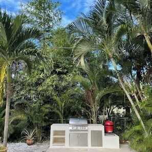 An outdoor kitchen with a grill and storage cabinets, surrounded by tropical vegetation.