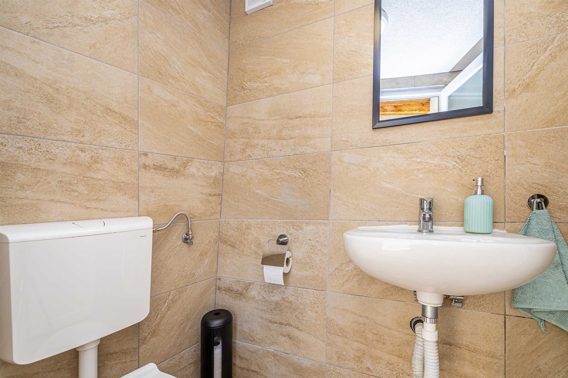 A bathroom with beige tiles, a white toilet, a sink with a faucet, a soap dispenser, and a towel hanging on a hook.