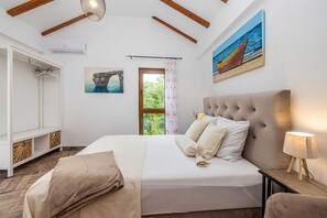A bedroom with a bed, bedside lamp, artwork, and a view of greenery. - Villa OASA with private heated pool (Ljutići)