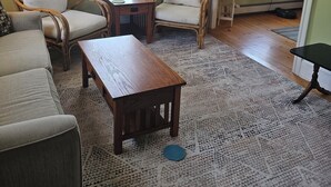 Ein Wohnzimmer mit einer Couch, zwei Sesseln, einem Couchtisch und einem Teppich. Es gibt zwei Fenster mit Jalousien.