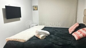 A bedroom with a bed, a TV mounted on the wall, a window with a blind, and a small bedside table. - Rbk Dpto new a pasos del Movistar Arena (Santiago)