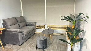 A modern living room with a grey sofa, a small round table, a grey ottoman, and a large green plant. - Rbk Dpto new a pasos del Movistar Arena (Santiago)