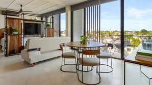 Ein modernes Wohnzimmer mit einer Couch, einem Esstisch, Stühlen, einem Fernseher und großen Fenstern mit Blick auf die Stadt.