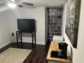 A room with a TV on a stand, a bookshelf filled with DVDs, a coffee maker on a table, and a ceiling fan.