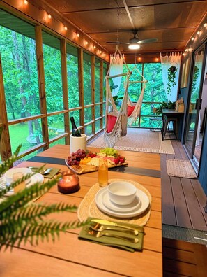 Ein Holztisch mit einer Obstplatte, einer Flasche und einem Glas Saft. Es gibt Hängelampen und einen Blick auf Bäume durch große Fenster.