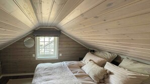 Une chambre avec un plafond en pente, un lit agrémenté de plusieurs oreillers, un miroir rond et une fenêtre donnant sur des immeubles.