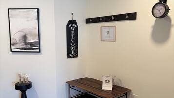Una habitación con piso de madera, una mesa negra con una bolsa de bienvenida blanca, un reloj de pared, un cartel de bienvenida en un marco y un cuadro abstracto enmarcado.
