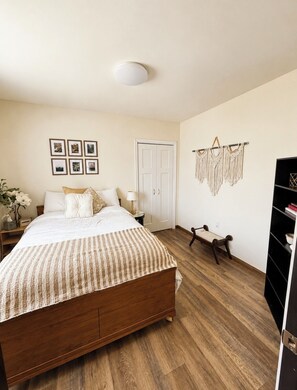 A bedroom with a wooden bed, a bedside table, a lamp, a bench, a wardrobe, and wall-mounted macramé decorations.
