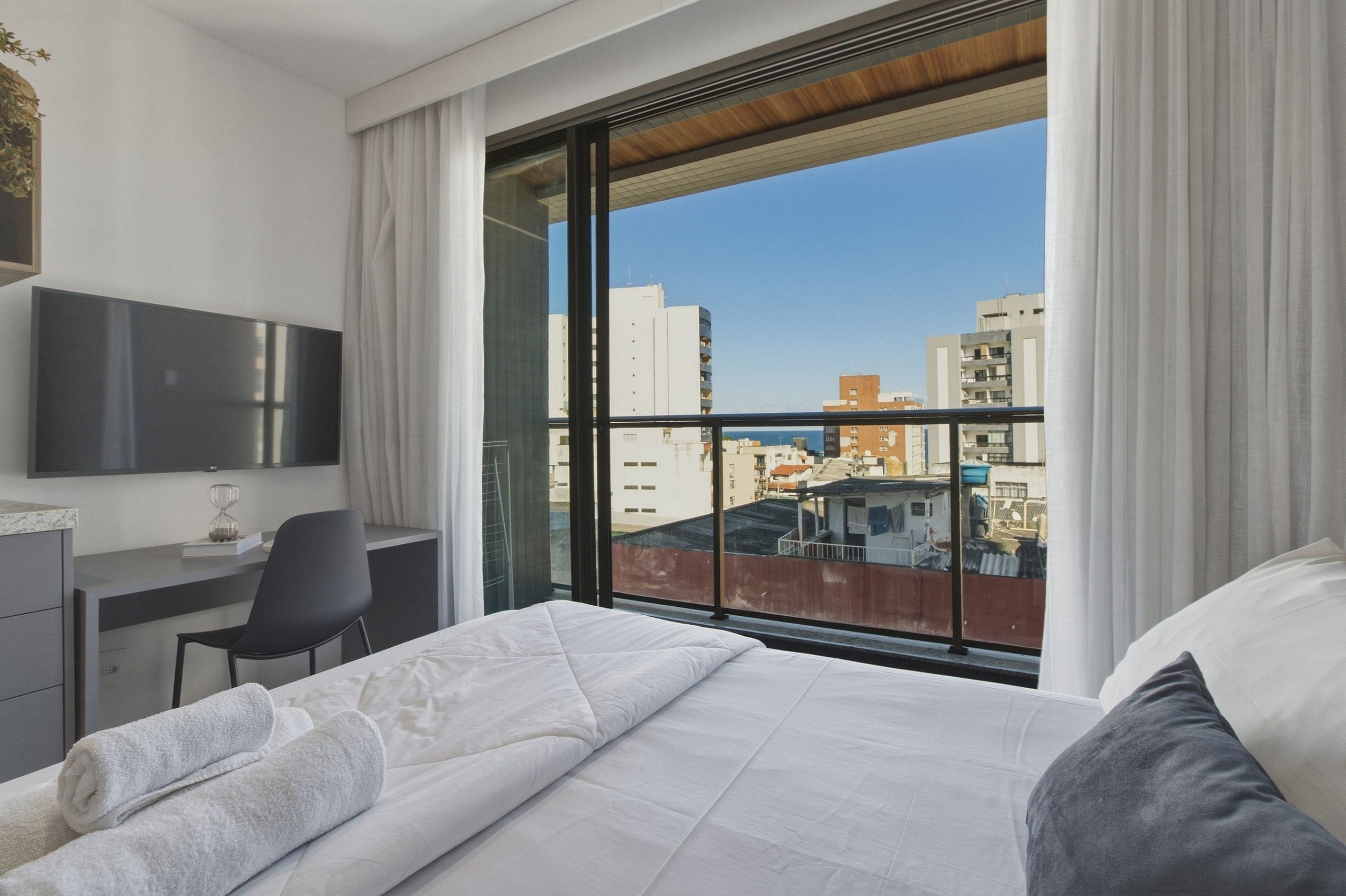 Ein modernes Hotelzimmer mit einem großen Bett, einem Schreibtisch, einem Sessel, einem Fernseher und einem Balkon mit Blick auf die Stadt.