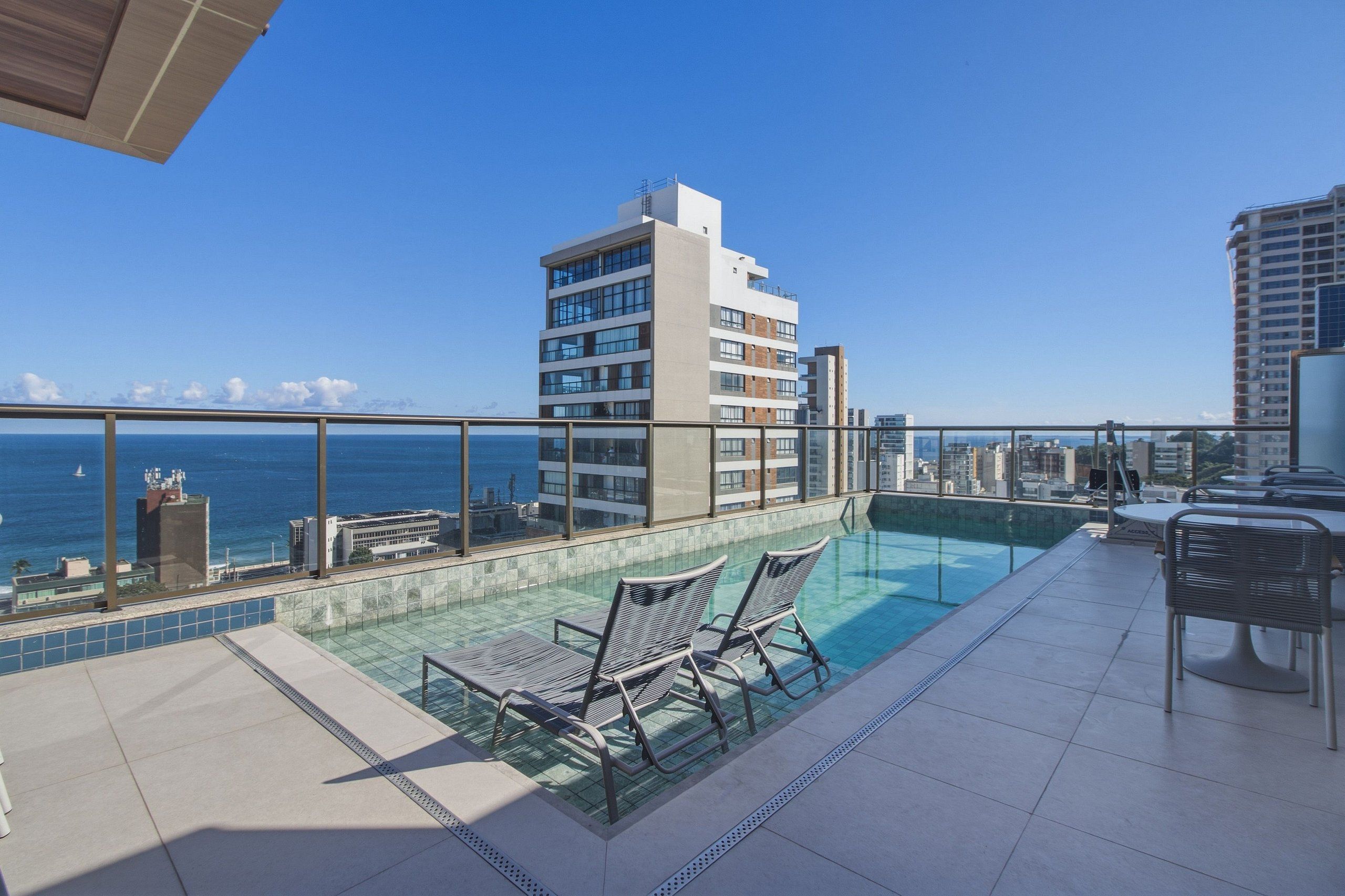 Una piscina en la azotea con dos sillas de descanso, una barandilla de vidrio y vistas al océano y al horizonte de la ciudad.