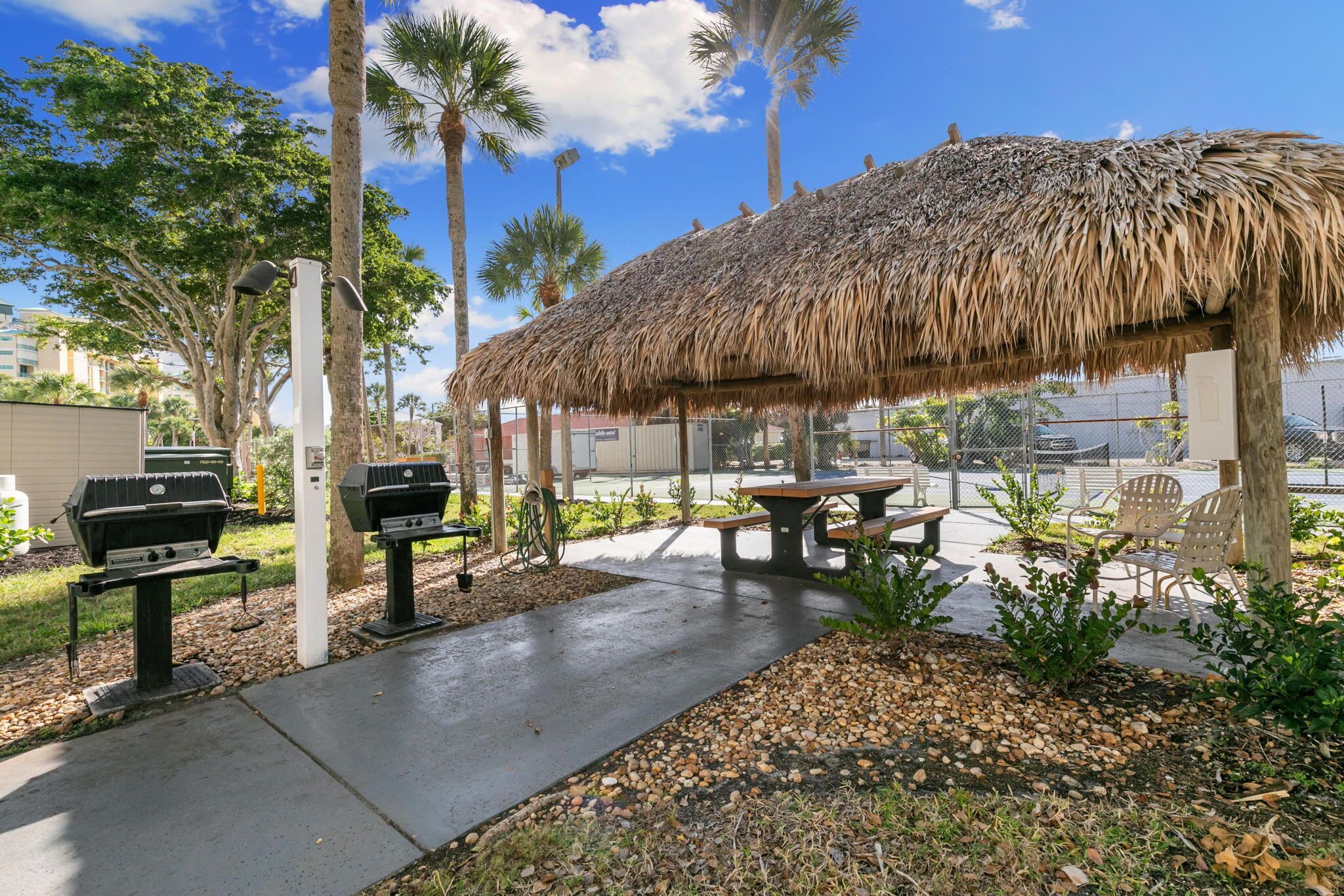 An outdoor patio with a thatched-roof structure, two barbecue grills, a picnic table, and chairs.