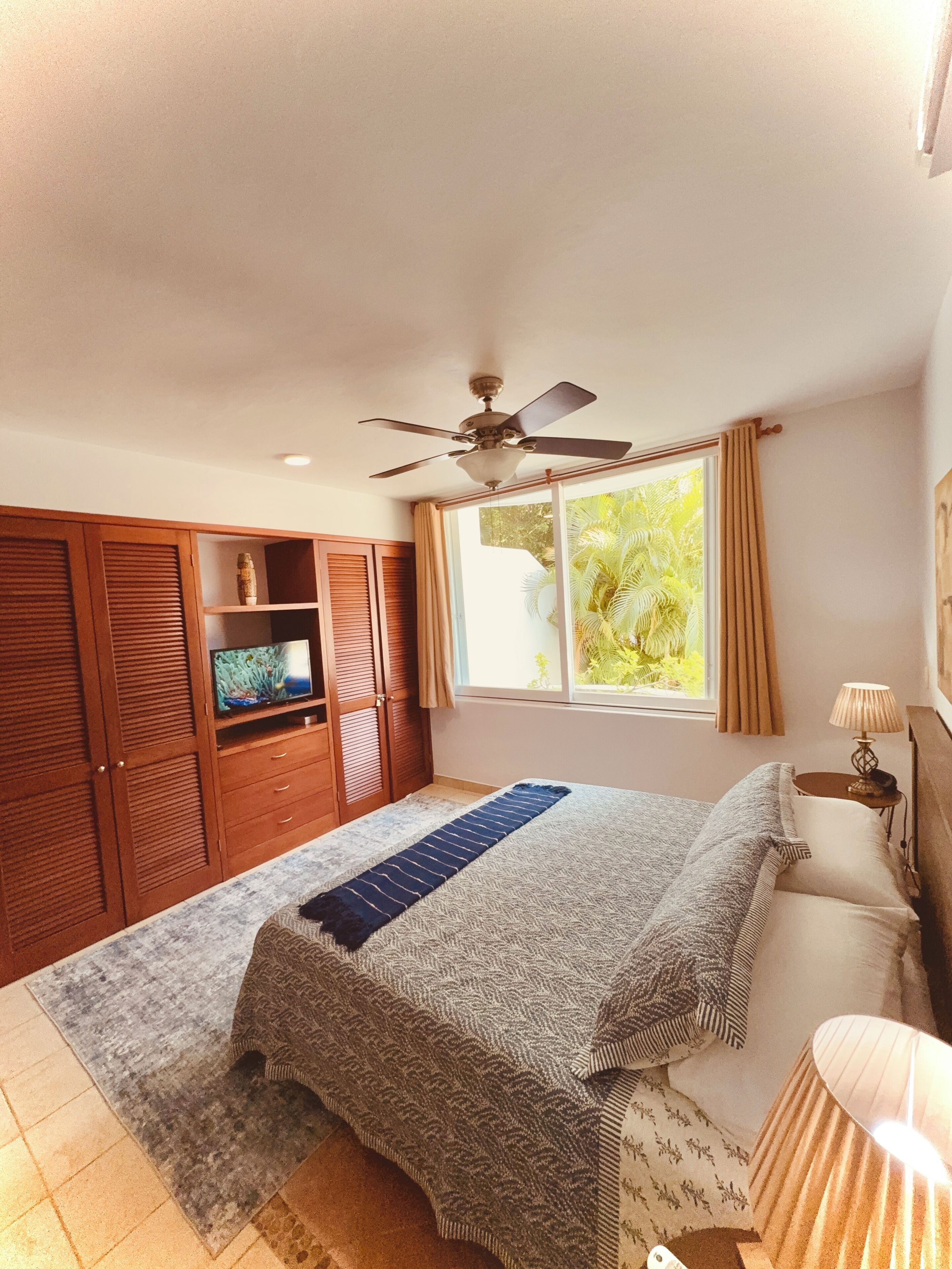 A bedroom with a bed, a ceiling fan, a TV on a wooden cabinet, a window with curtains, and a bedside lamp.