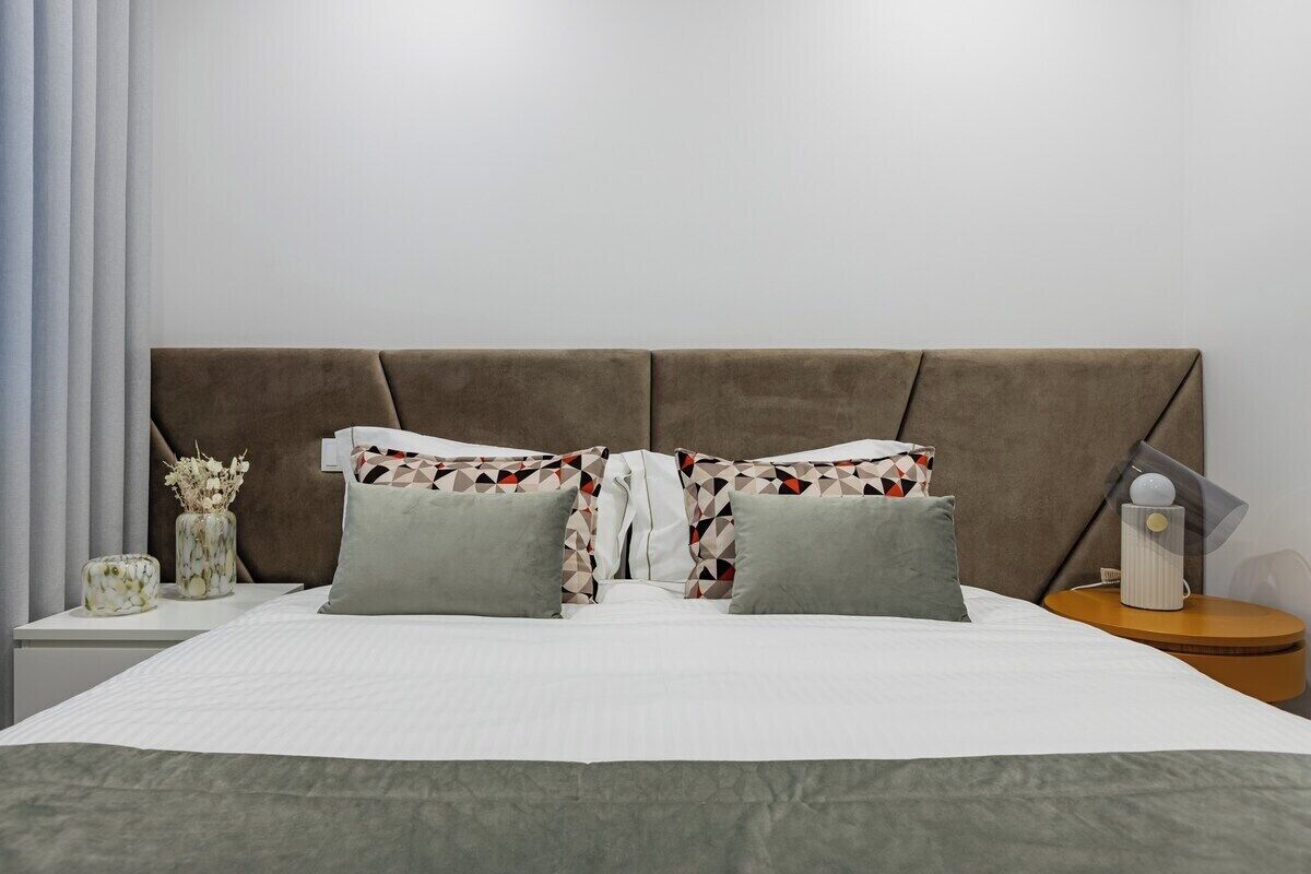Een bed met hoofdbord, kussens, nachtkastje en een vaas met gedroogde bloemen.