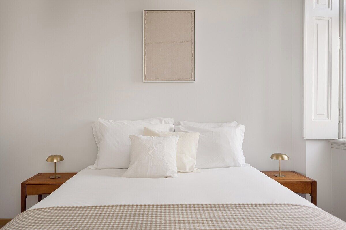 Een netjes opgemaakt bed met witte beddengoed en een geruit plaid, twee bedlampen en een ingelijste foto aan de muur.