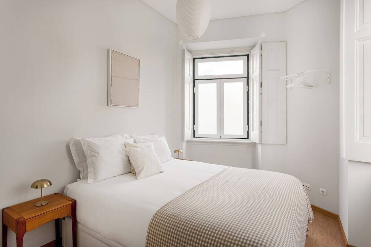 Ein Schlafzimmer mit Bett, Nachttisch, Lampe, Fenster und Wandleuchte.