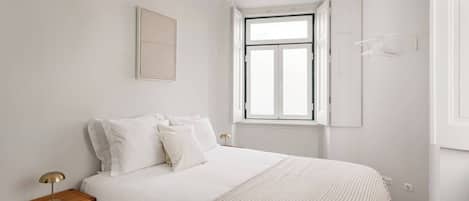 Un dormitorio con cama, mesita de noche, lámpara, ventana y aplique en la pared.